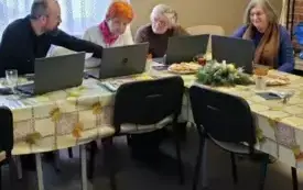 Na rogu stołu z kolorową ceratą siedzi grupa 4 os&oacute;b, w tym 3 kobiety, seniorki oraz 1 mężczyzna z brodą, instruktor. Seniorki wpatrzone są w otwarte przed sobą laptopy. Mężczyzna ma przed sobą otwarty laptop i patrzy w stronę seniorek coś im tłumacząc. Na