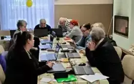 Grupa 9 senior&oacute;w, w tym 7 kobiet, 2 mężczyzn, siedzących na krzesłach przy długim stole z kolorową ceratą. Na stole stoi 9 otwartych laptop&oacute;w, w kt&oacute;re patrzą seniorzy. Naprzeciw widać okno z białą firanką udekorowaną kolorowymi balonami oraz niebiesko bia