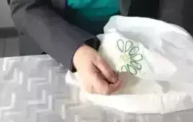 Pani w zielonym golfie i szarej marynarce siedzi na krześle przy stole na kt&oacute;rym jest srebrna cerata. Trzyma w ręce poszewkę w kolorze ecru na kt&oacute;rej haftuje kwiat. Wzorem łańcuszkowym zrobiła płatki a w środku ż&oacute;łte k&oacute;łko. Pani z lekko opuszczonymi okula