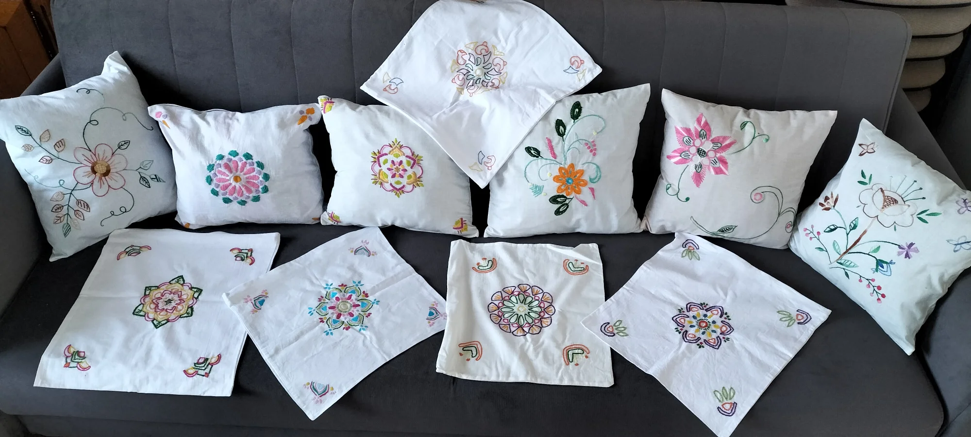 Szare ł&oacute;żko na&nbsp;kt&oacute;rym ułożone są haftowane poduszki. Część poduszek ustawiona jest w&nbsp;rzędzie przy oparciu ł&oacute;żka a&nbsp;reszta poszewek ułożona przed nimi z&nbsp;przodu na&nbsp;siedzisku. Każda poduszka przedstawia inny wz&oacute;r i&nbsp;kolor. Widoczne są cztery poduszki, kt&oacute;re przedstawiają kolorowe kwiaty, łodygi i&nbsp;zielone liście. Na jednej z&nbsp;nich widać niebieskie motyle.